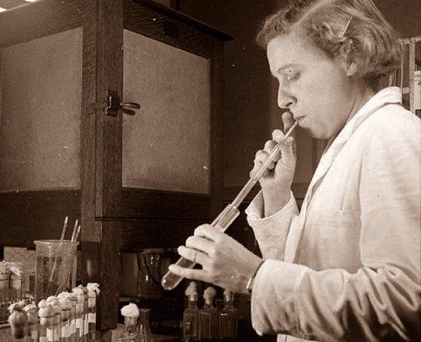 Imagen en blanco y negro de una mujer en el laboratorio usando la boca para pipetear células de un tubo de ensayo a otro.